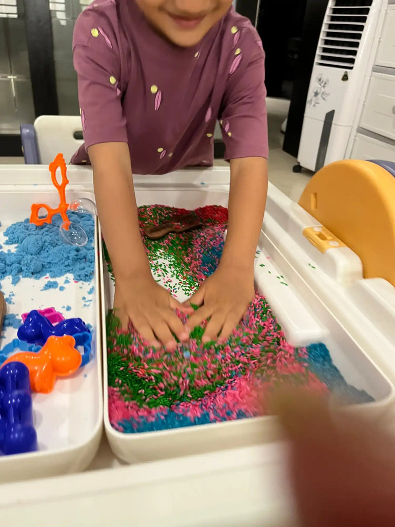 A child’s sensory play table with two bins, one containing blue kinetic sand with toys and the other filled with colorful rice for tactile exploration.