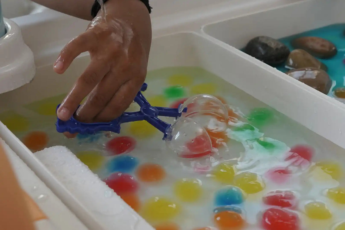 A hand holding a blue scoop filled with colorful water beads.