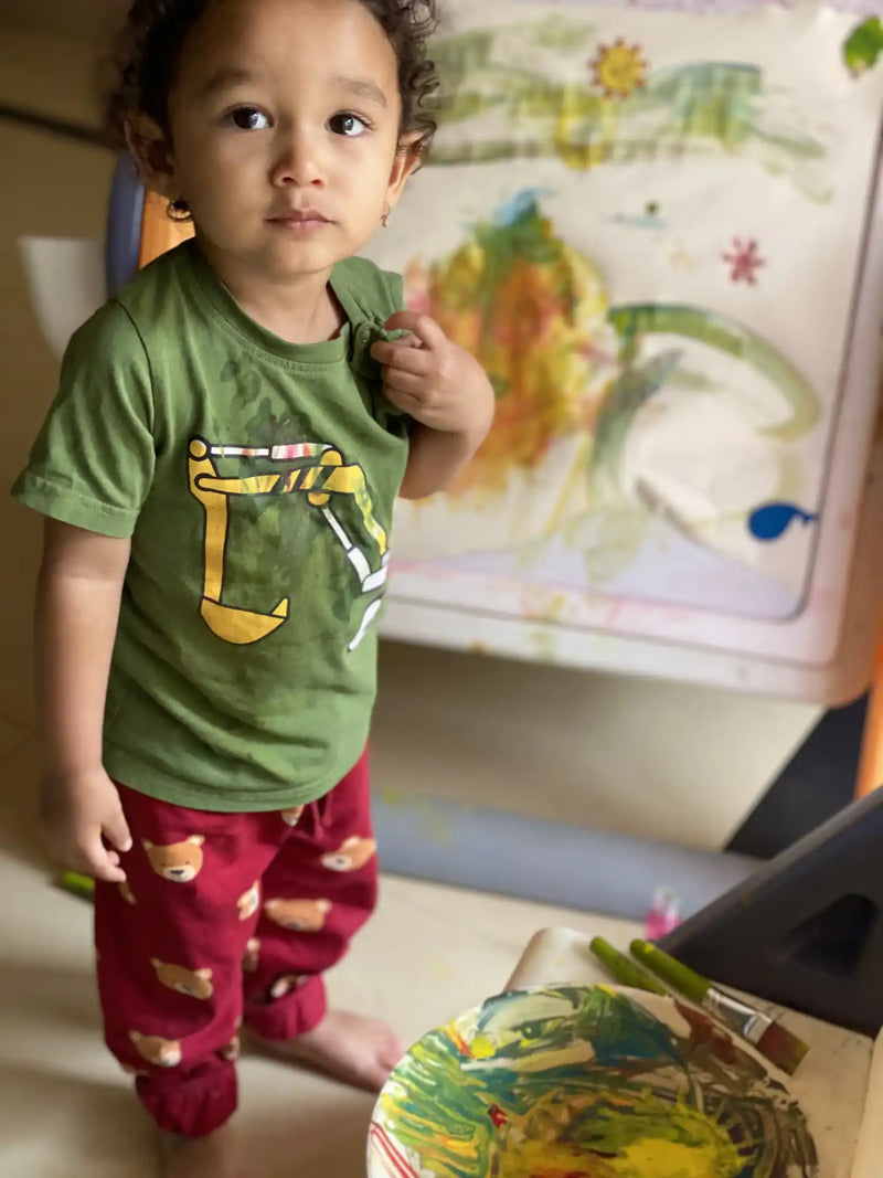 A toddler wearing a green shirt with a yellow excavator design and red pants with bear patterns, standing barefoot next to an easel with a colorful painting and a paint palette on the floor.