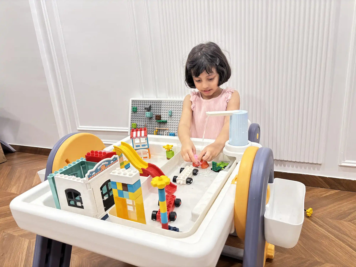 A young child playing with a multi-functional activity table featuring building blocks, a slide, and toy cars.