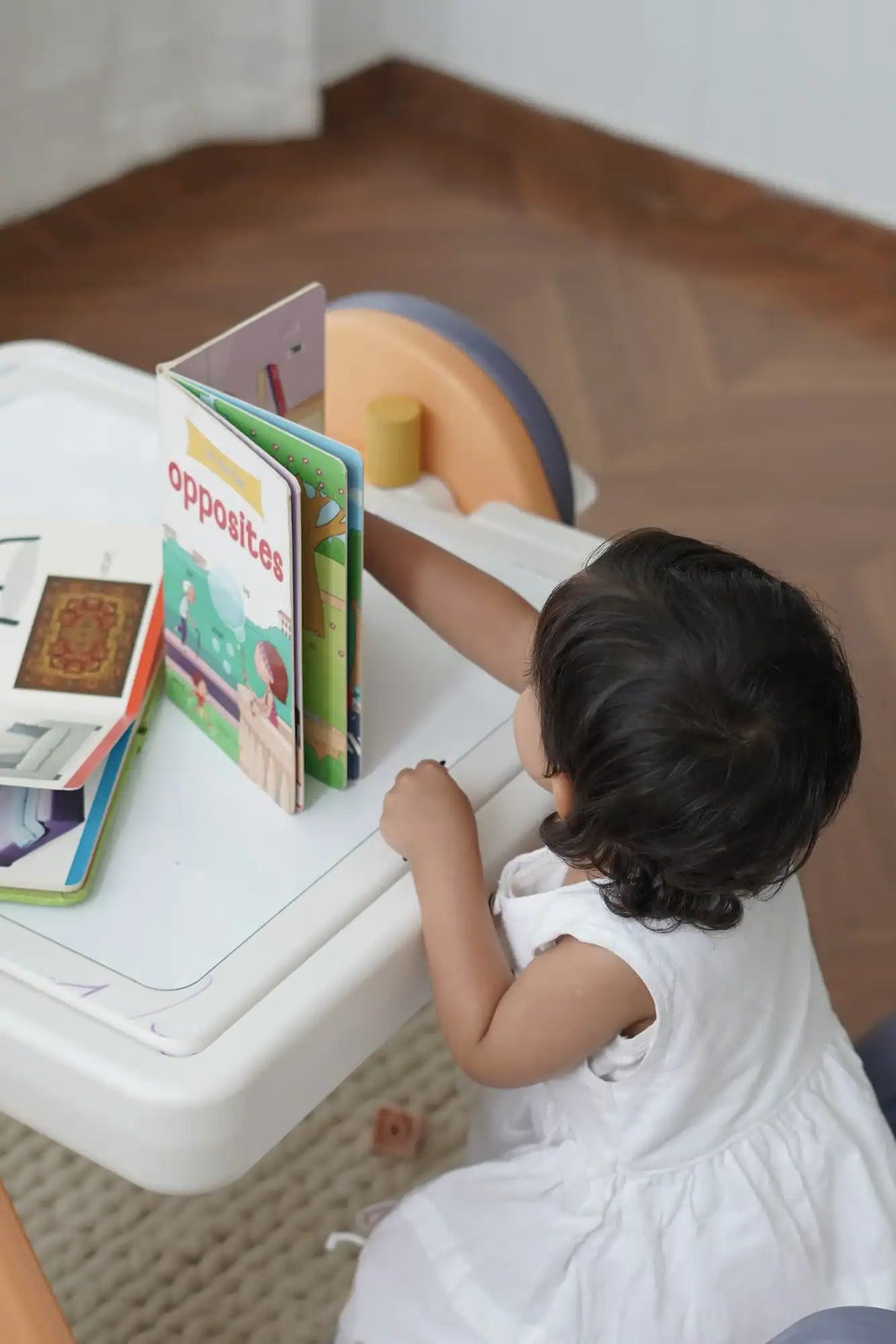 A young child in a white dress interacting with a children’s book titled ’opposites’ on a white table.